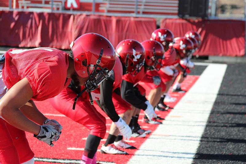 Photo by: Josh Friesen

Cornerbacks and safeties prepare in athletic stances to begin a drill during practice. 