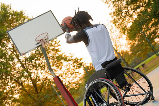 Young athlete on wheelchair throwing ball in basketball basket, sunset effect in background.